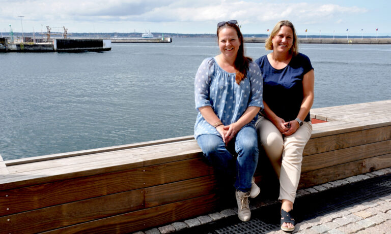 Två kvinnor sitter vid en kajkant med hav, färja och flaggor i bakgrunden.